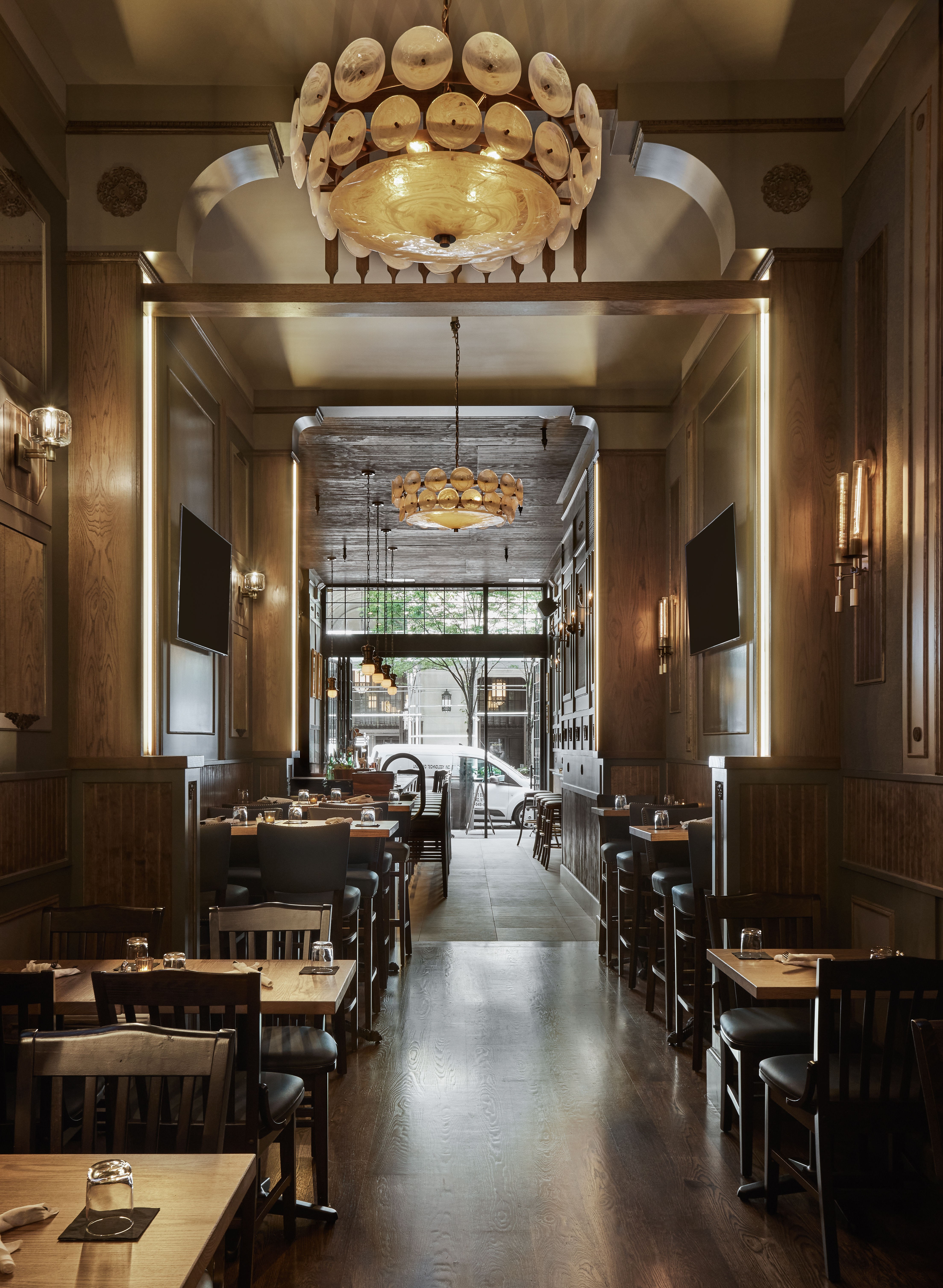 Cozy upscale wood-paneled downtown restaurant interior with warm shell chandeliers, polished dark hardwood floors, rows of wooden dining tables and bar seating, and a street-facing window with trees.