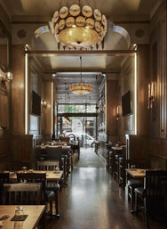 Cozy upscale wood-paneled downtown restaurant interior with warm shell chandeliers, polished dark hardwood floors, rows of wooden dining tables and bar seating, and a street-facing window with trees.