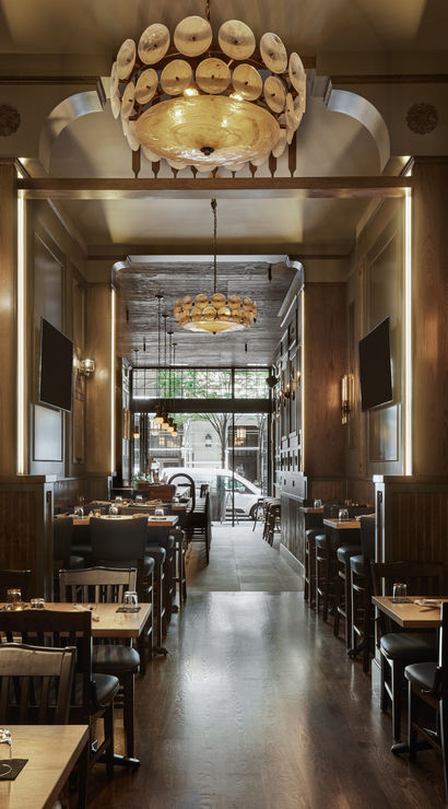 Cozy upscale wood-paneled downtown restaurant interior with warm shell chandeliers, polished dark hardwood floors, rows of wooden dining tables and bar seating, and a street-facing window with trees.