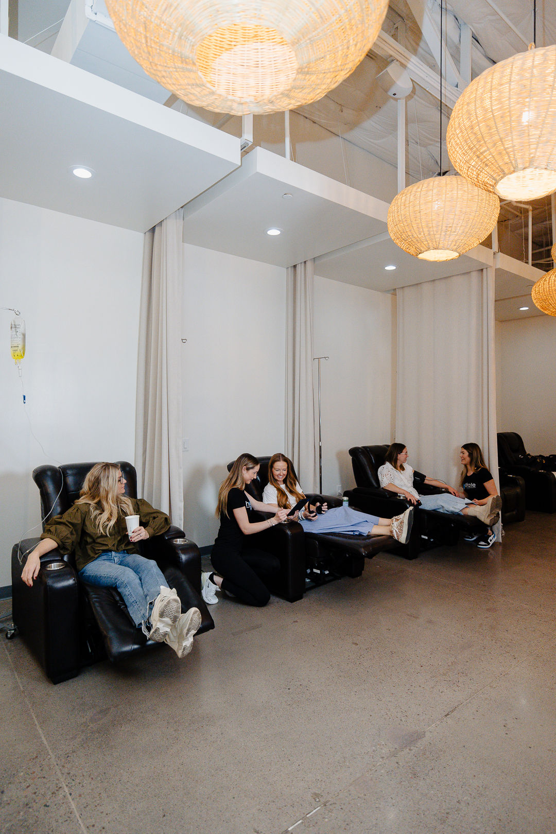 Spa-like IV therapy lounge with wicker pendant lights, white curtains and several people relaxing in black reclining chairs while receiving IV hydration in a bright, modern wellness studio.