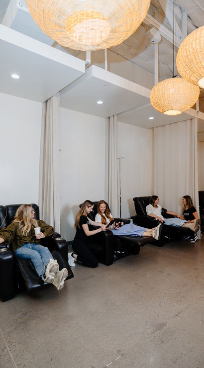 Spa-like IV therapy lounge with wicker pendant lights, white curtains and several people relaxing in black reclining chairs while receiving IV hydration in a bright, modern wellness studio.