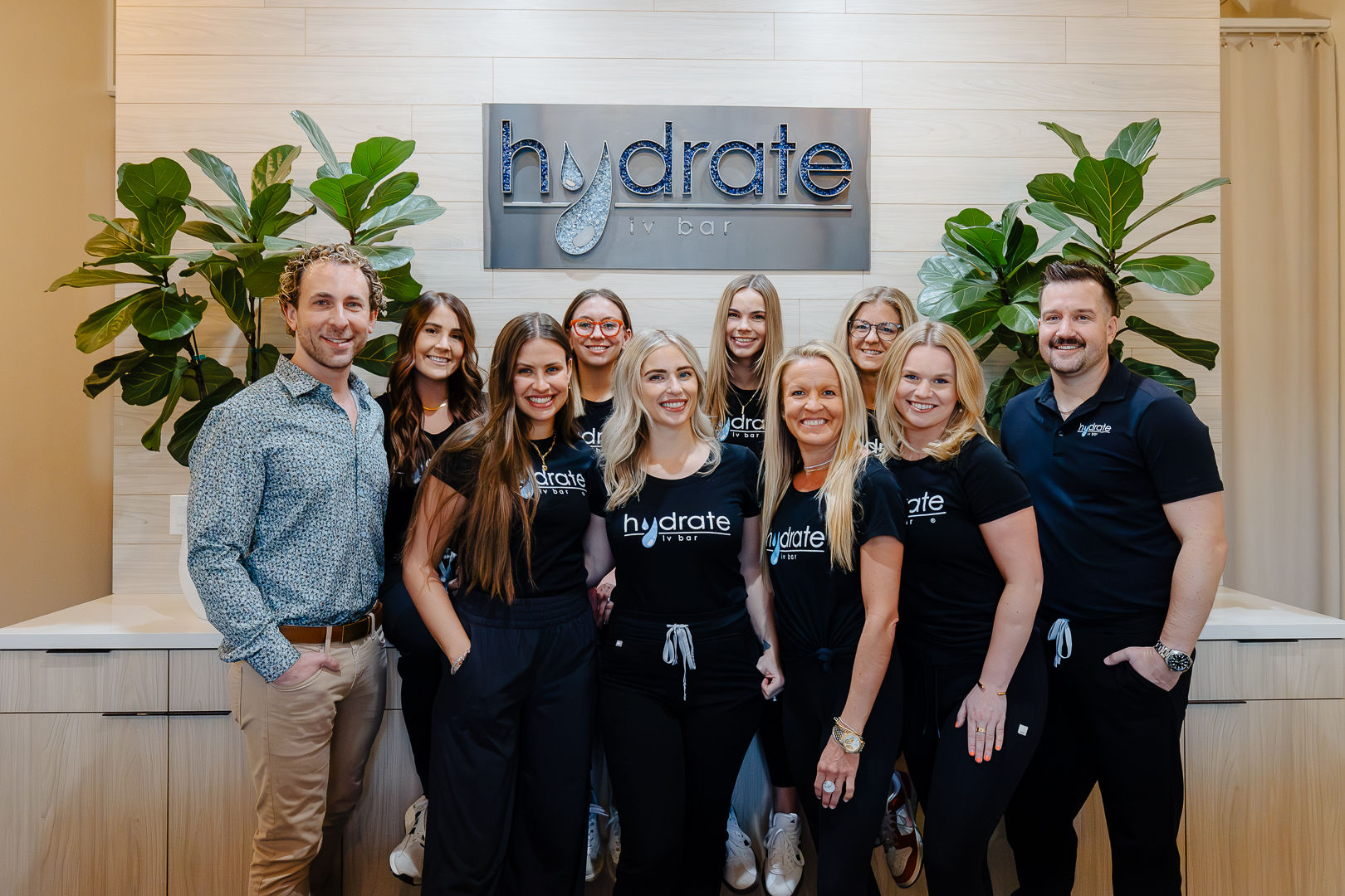Smiling IV hydration clinic team – eleven staff in matching black shirts and two men posing in a bright, modern reception with wood-paneled wall and large green plants.
