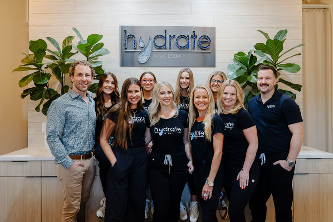 Smiling IV hydration clinic team – eleven staff in matching black shirts and two men posing in a bright, modern reception with wood-paneled wall and large green plants.