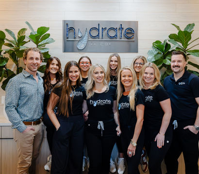 Smiling IV hydration clinic team – eleven staff in matching black shirts and two men posing in a bright, modern reception with wood-paneled wall and large green plants.