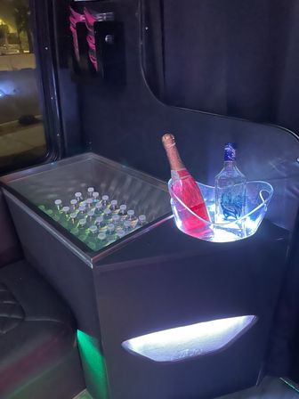 LED-lit party bus interior with a built-in cooler of bottled water and an illuminated ice bucket holding pink champagne and a clear liquor bottle, ready for celebration