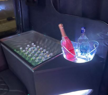 LED-lit party bus interior with a built-in cooler of bottled water and an illuminated ice bucket holding pink champagne and a clear liquor bottle, ready for celebration