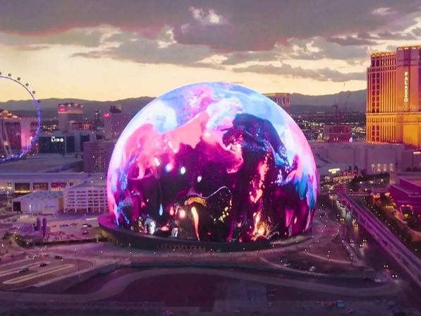 Giant illuminated digital sphere displaying swirling pink and blue visuals on the Las Vegas skyline at dusk, surrounded by city lights, a Ferris wheel, and nearby high-rise buildings.