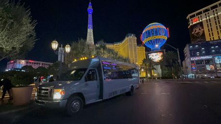 Las Vegas Strip at night: white passenger shuttle parked beneath illuminated Eiffel-style tower and colorful neon balloon sign