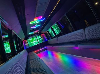 Vibrant party bus interior with quilted leather bench seating, center dance pole, wood-look floor and multicolored LED ceiling and floor lights reflecting on windows.