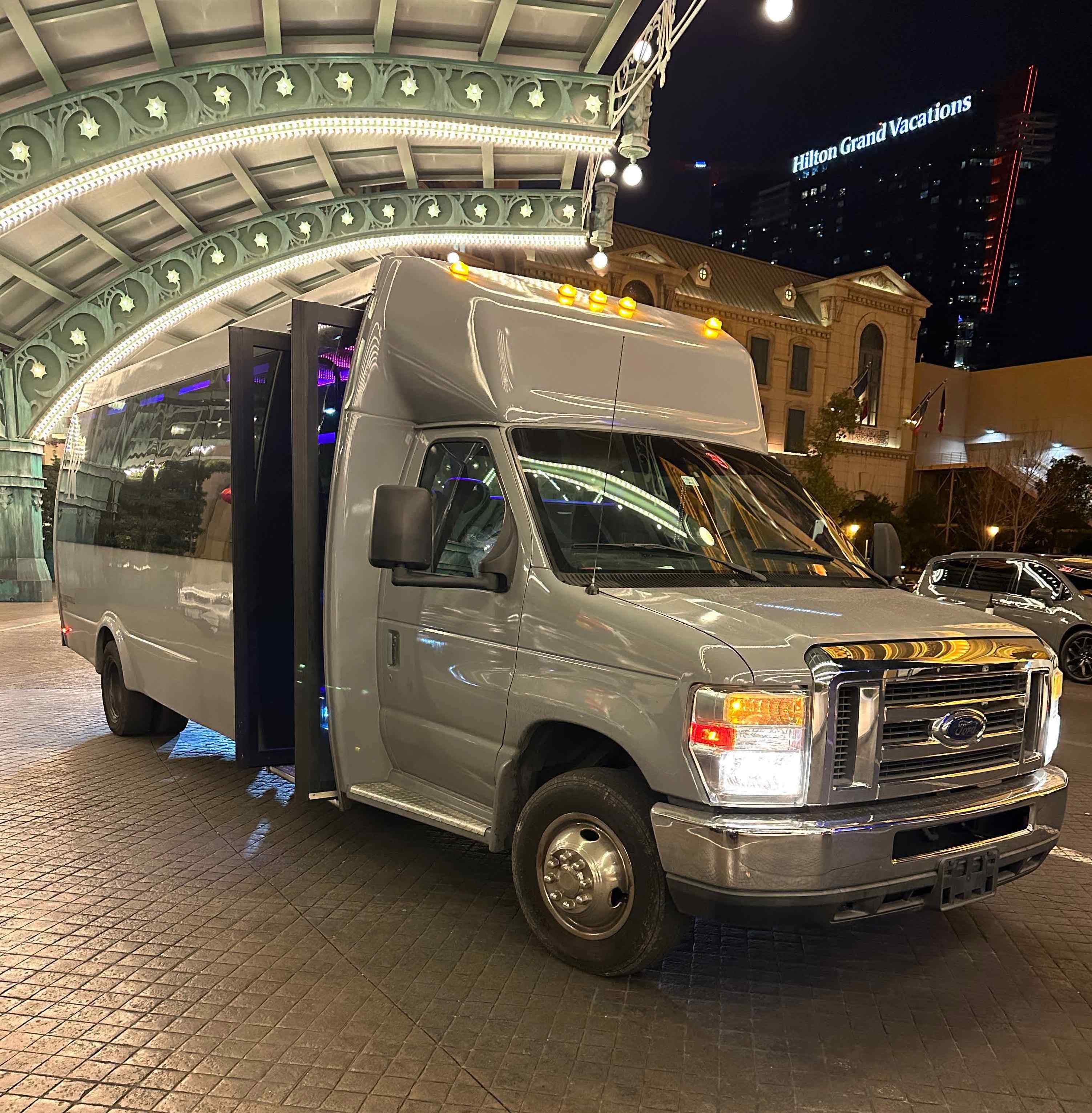 Silver passenger shuttle van with open door parked under an ornate illuminated hotel canopy at night outside a Las Vegas hotel, city lights and skyline reflected on the vehicle