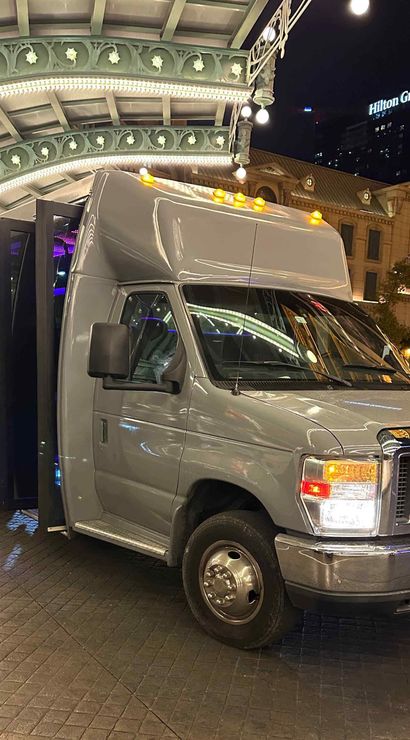 Silver passenger shuttle van with open door parked under an ornate illuminated hotel canopy at night outside a Las Vegas hotel, city lights and skyline reflected on the vehicle