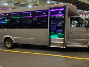 Luxury party shuttle van parked at an airport terminal curb at night, open door revealing purple and green LED-lit interior with leather bench seats and entry steps.
