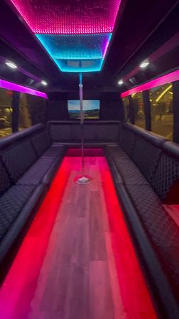 Nighttime party bus interior with U-shaped black leather seating, center dance pole, wood-look floor and vibrant pink and blue LED ceiling and floor lighting.