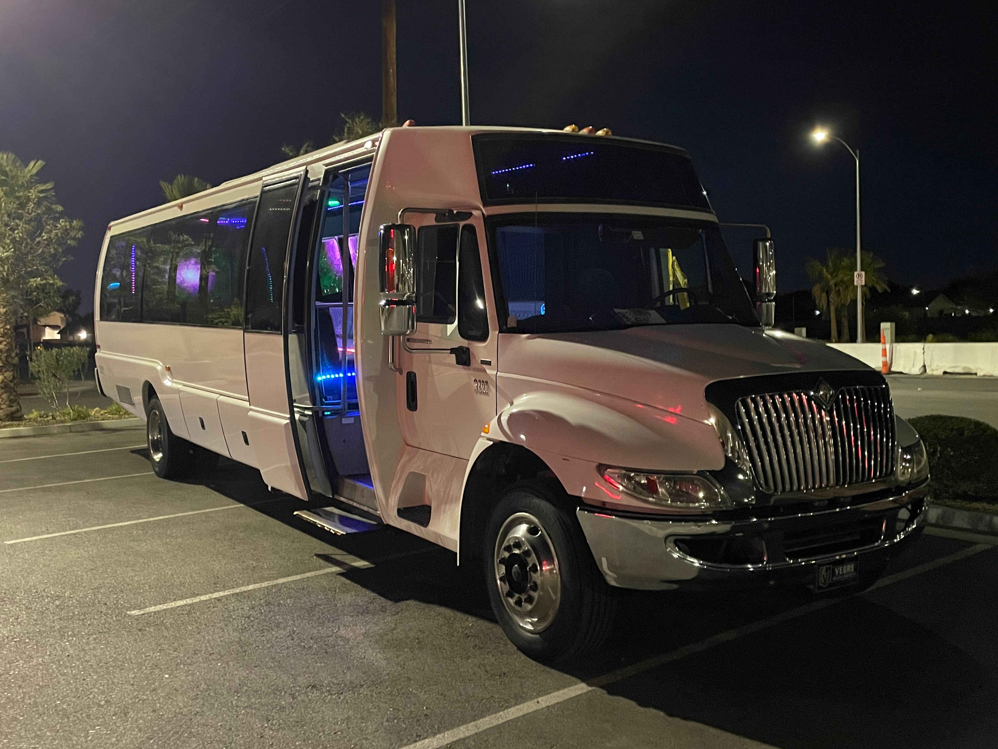 White party bus parked in a dimly lit parking lot at night, front door open to show blue and purple LED interior lights and chrome grille reflecting streetlight