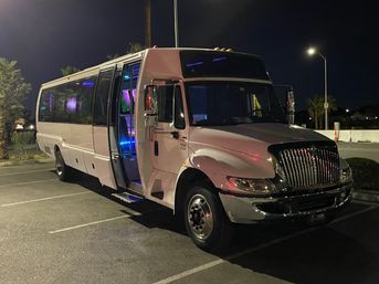 White party bus parked in a dimly lit parking lot at night, front door open to show blue and purple LED interior lights and chrome grille reflecting streetlight