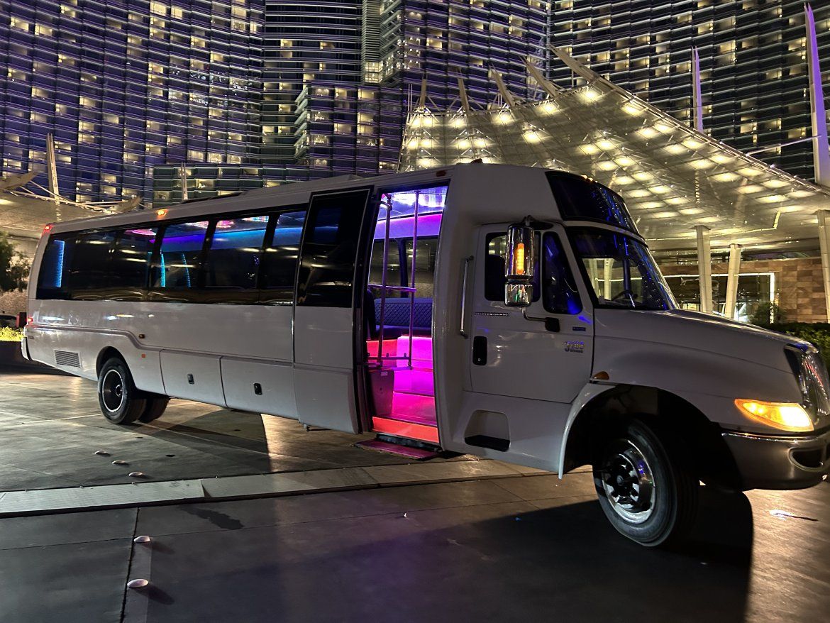 White party bus with open door revealing pink and purple LED interior lights parked at a luxury hotel entrance beneath illuminated high-rise glass facade at night