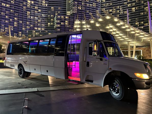 White party bus with open door revealing pink and purple LED interior lights parked at a luxury hotel entrance beneath illuminated high-rise glass facade at night
