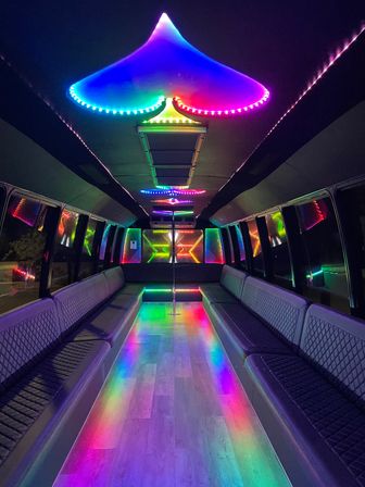 Vibrant party bus interior with rainbow LED canopy and strip lights, mirrored panels, center dance pole and wood-look floor reflecting multicolor neon, quilted bench seating along both sides.