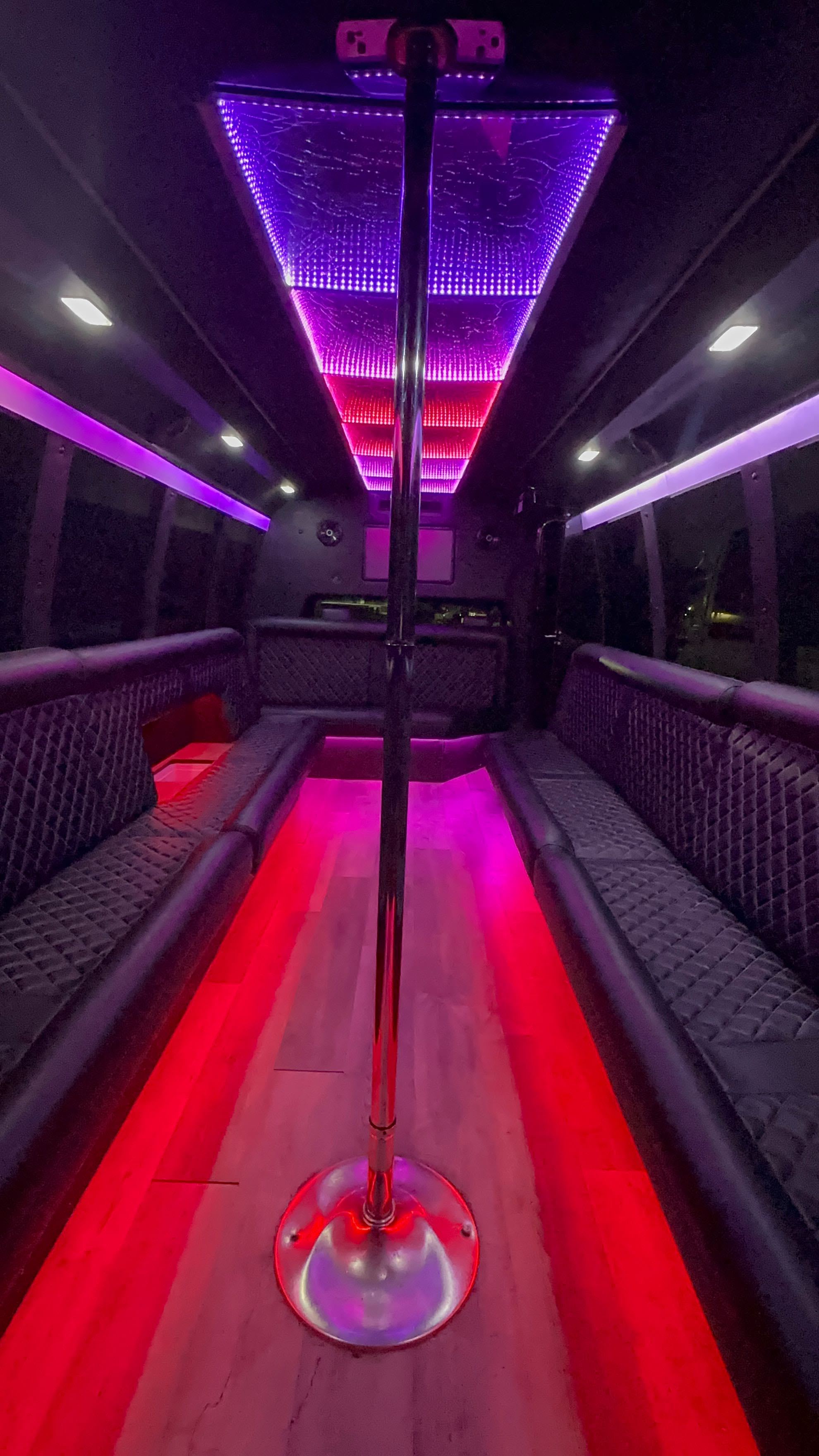 Vibrant party bus interior with center chrome dance pole, purple and red LED ceiling and floor lighting, quilted black leather bench seating and wood-look flooring