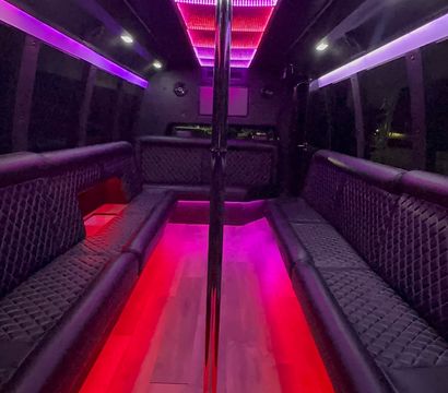Vibrant party bus interior with center chrome dance pole, purple and red LED ceiling and floor lighting, quilted black leather bench seating and wood-look flooring