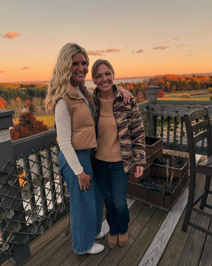 Two smiling women posing on a wooden deck at sunset, wearing cozy fall layers with colorful autumn trees and a distant lake view
