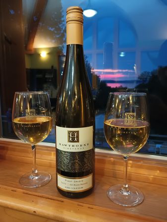 Pinot Grigio bottle flanked by two filled wine glasses on a wooden windowsill, warm indoor light and a pink‑blue lakeside sunset reflected in the window.