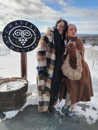 Two friends in cozy winter coats posing by a decorative round winery sign, snow-covered vineyard and rolling hills under a bright blue winter sky