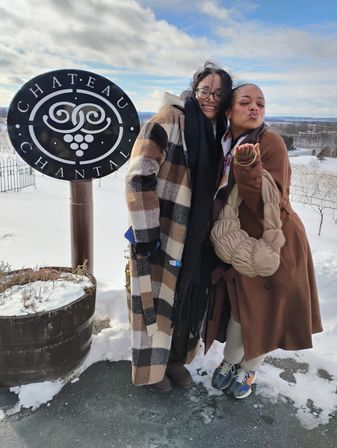 Two friends in cozy winter coats posing by a decorative round winery sign, snow-covered vineyard and rolling hills under a bright blue winter sky