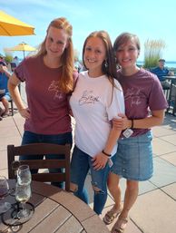 Smiling bride in a white "Bride" tee with two friends in matching maroon "Bride Tribe" shirts on a sunny lakeside patio with yellow umbrellas and wine glasses on a table.