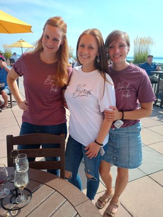 Smiling bride in a white "Bride" tee with two friends in matching maroon "Bride Tribe" shirts on a sunny lakeside patio with yellow umbrellas and wine glasses on a table.