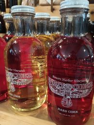 Close-up of glass bottles of red and golden hard cider with decorative white labels and white caps lined up on a wooden shelf
