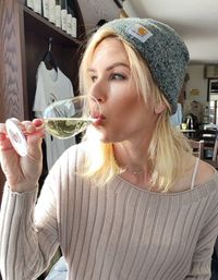 Blonde woman in a gray beanie and ribbed sweater sipping white wine from a stemmed glass in a cozy winery tasting room