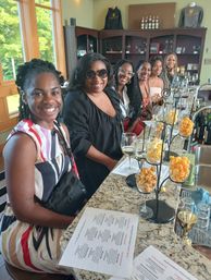 Cheerful group of six friends seated at a sunlit winery tasting room counter with wine glasses, menus and popcorn-pairing displays on a granite bar.
