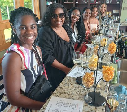 Cheerful group of six friends seated at a sunlit winery tasting room counter with wine glasses, menus and popcorn-pairing displays on a granite bar.