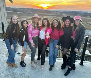 Seven friends in pink and black outfits posing on a rooftop terrace overlooking a vineyard at sunset with rolling fields and a golden sky.