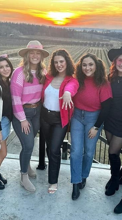 Seven friends in pink and black outfits posing on a rooftop terrace overlooking a vineyard at sunset with rolling fields and a golden sky.