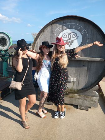 Three friends wearing cowboy hats posing on a sunny winery patio in front of a large wooden wine barrel, with a lake view and blue sky in the background.