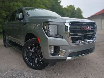 Sleek silver GMC SUV front three-quarter view parked on a gravel driveway, chrome grille, LED headlight and black alloy wheel with trees in the background