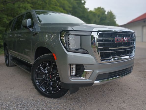 Sleek silver GMC SUV front three-quarter view parked on a gravel driveway, chrome grille, LED headlight and black alloy wheel with trees in the background