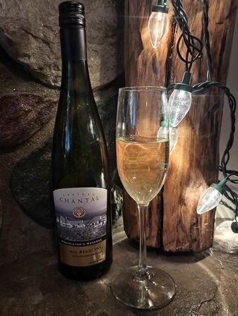 Riesling wine bottle next to a filled white wine flute on a stone countertop, warm string lights wrapped around rustic wood for a cozy, rustic tasting-room vibe
