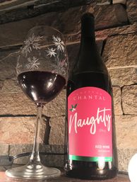 Festive Michigan red wine bottle with bright pink "Naughty" label and holly accent beside a stemmed wine glass etched with snowflake designs, both on a stone ledge.