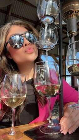 Person in sunglasses and pink jacket puckering for a selfie beside a stacked wine-tasting flight of white wine on a sunny outdoor patio.