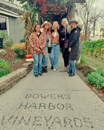 Five women smiling on a vineyard walkway under a pergola draped with autumn vines, pumpkins on a stone border and grapevines and bare trees in the background.