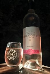 Chilled rosé wine bottle and logo-etched stemless glass glowing on a wooden deck railing at night on Old Mission Peninsula, Michigan
