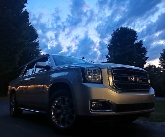 Silver full-size SUV with gleaming chrome grille parked on a suburban driveway at dusk, low-angle shot against a dramatic blue cloudy sky and tree silhouettes.