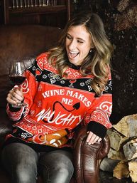 Laughing woman in a red holiday sweater reading "Wine Makes Me Naughty" holding a glass of red wine while seated on a leather armchair next to stacked firewood