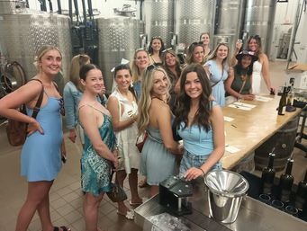 Smiling group of women in summer dresses enjoying a wine tasting inside a winery production room with stainless steel fermentation tanks and a long wooden tasting table with bottles.