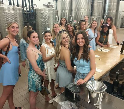Smiling group of women in summer dresses enjoying a wine tasting inside a winery production room with stainless steel fermentation tanks and a long wooden tasting table with bottles.