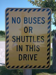 Yellow roadside sign with black diagonal border reading “NO BUSES OR SHUTTLES IN THIS DRIVE,” posted at a driveway entrance in a park-like setting.