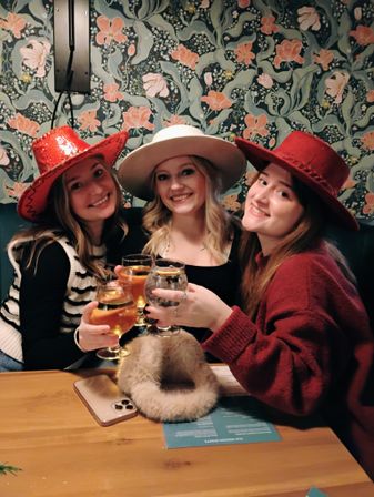 Three friends in festive hats smiling and clinking beer and cocktail glasses at a cozy floral-wallpapered cocktail bar booth